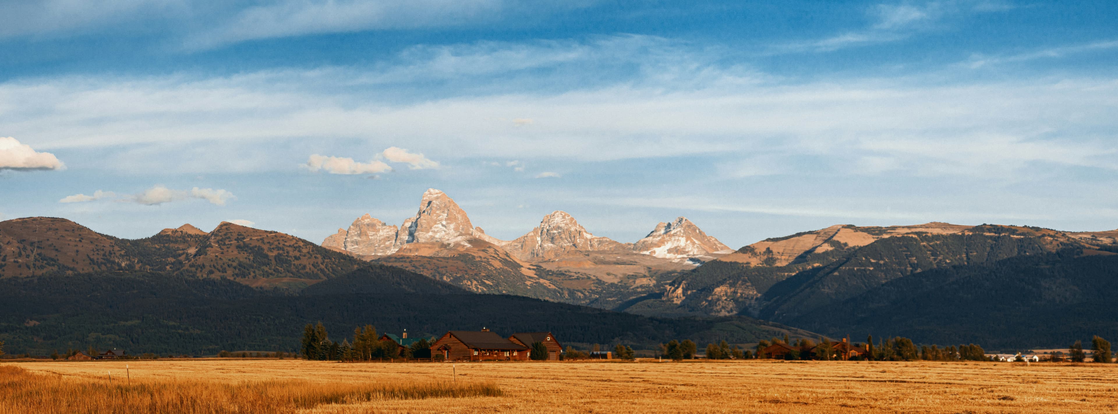 Ranch buildings at the base of the Teton range with golden fields in the foreground